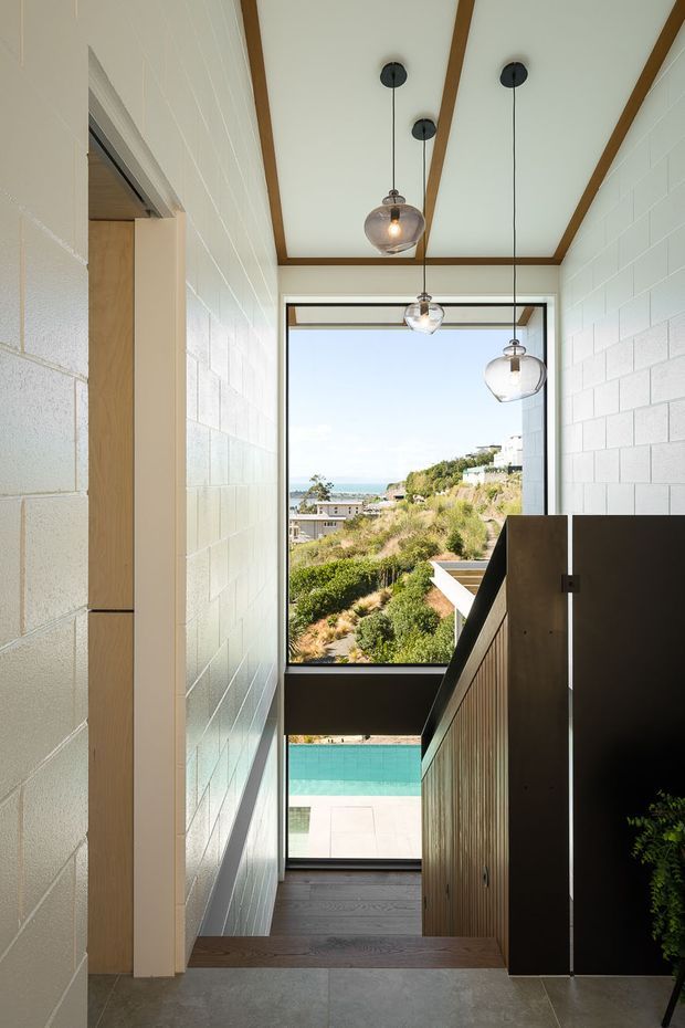 A bright, narrow interior stairway or landing with white-painted block walls, exposed timber ceiling beams, dark timber flooring, and contemporary glass pendant lights. A timber-and-dark-panel stair balustrade descends toward large windows overlooking a landscaped hillside, ocean or water beyond, and a turquoise swimming pool. The space has strong vertical proportions and abundant natural light.