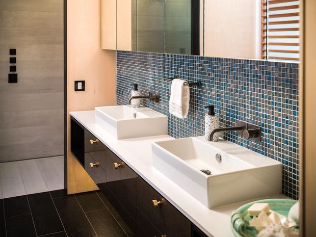 Contemporary bathroom vanity with two white vessel basins, a long white benchtop, dark timber cabinetry with brass pulls, and wall-mounted brushed metal tapware. The feature wall is clad in small blue-grey mosaic tiles, with pale timber cabinetry above and a large mirror. Dark floor tiles, a towel warmer, and warm natural lighting complete the refined double-vanity arrangement.