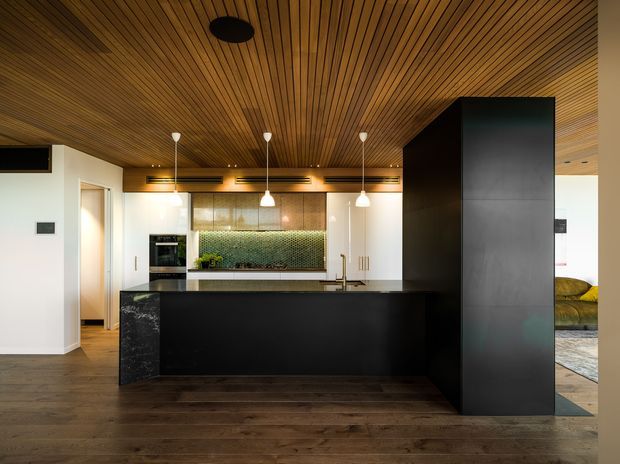 A contemporary kitchen viewed straight on, featuring a large matte-black island with a dark stone-look end panel, white cabinetry, integrated appliances, brass-toned hardware and tapware, and a green mosaic-tile backsplash. A full-height dark storage wall partially screens an adjacent living area. The room is defined by a warm slatted timber ceiling, dark timber flooring, and three pendant lights over the island.