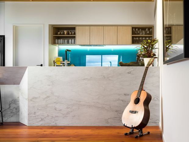 Contemporary kitchen viewed from the living area, featuring a large pale marble-look island with waterfall ends, warm timber flooring, light timber overhead cabinetry, open shelving with glassware, and a vivid turquoise glass splashback. A guitar rests against the island, while a glazed opening and indoor plants add visual interest.