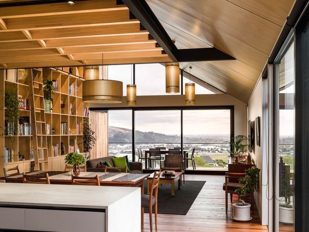A warm, light-filled open-plan living and dining area with a partially visible contemporary kitchen island in the foreground. The room features extensive floor-to-ceiling glazing overlooking a distant hillside and urban landscape, a dramatic sloped timber-lined ceiling with exposed dark structural beams, exposed timber ceiling battens, pendant lights, timber flooring, a large built-in shelving wall, dining table, and lounge seating. The palette is predominantly warm timber, white, grey, and black, creating a refined yet inviting atmosphere.