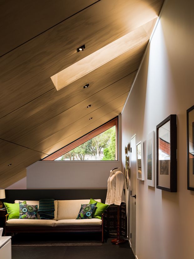 A compact, warmly lit living room with a dramatic sloping timber-lined ceiling and clerestory-style triangular window. A cream sofa with colourful cushions sits against a dark low wall, while framed artwork and a coat rack line the pale side wall. Recessed spotlights and a long rooflight-like ceiling opening emphasize the angular architecture.