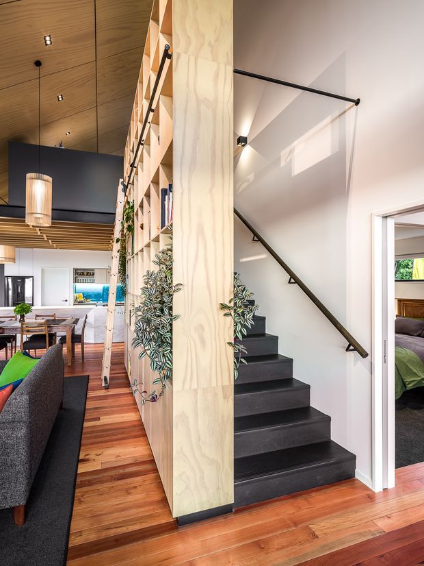 Interior view of a contemporary open-plan home centred on a staircase with dark carpeted treads, a slender black metal handrail, and a tall plywood-lined partition with open display shelving and climbing plants. Warm timber flooring and ceiling panelling continue through the dining and living area, with a bedroom visible through a doorway at right. The space has strong vertical joinery, integrated lighting, and a bright, open feel.