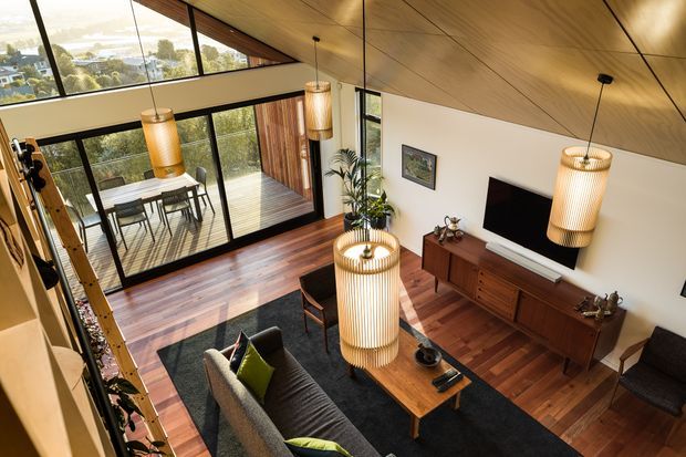A warm, double-height living room with a dramatic timber-lined sloping ceiling, extensive black-framed glazing, and an adjoining outdoor deck. The room contains a grey sofa, dark rug, timber coffee table, mid-century-style timber media console, television, armchair, and several large ribbed pendant lights. A dining area is visible through the glazed opening, with leafy suburban views beyond.
