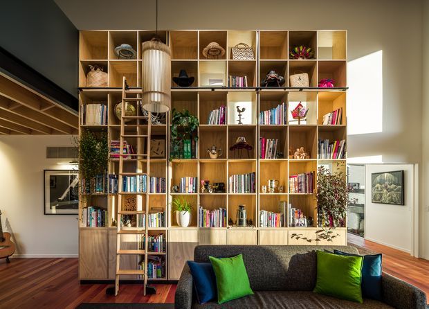 A bright, double-height living room dominated by a large custom timber open shelving and cabinetry wall filled with books, decorative objects, plants, and artwork. A timber ladder provides access to the upper shelves, while a pendant light hangs in front of the joinery. A dark sofa with vivid green and blue cushions sits in the foreground. Warm timber flooring, white walls, strong daylight, and visible upper-level architectural elements create a contemporary, inviting interior.
