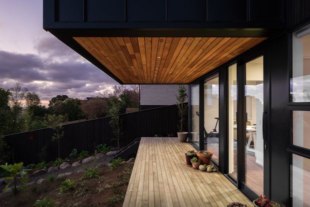 A contemporary covered timber deck beside a dark-clad house, with full-height glazed doors opening to a bright interior. The deck extends over a landscaped garden and is framed by a projecting upper structure with a warm timber-lined soffit. The image is photographed at dusk, creating a dramatic contrast between the black exterior, amber timber, and cloudy sky.