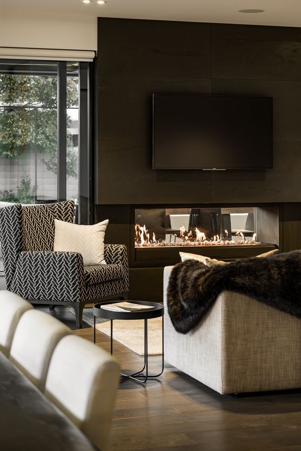 A sophisticated living room with a wall-mounted television above a long linear fireplace set into a dark paneled feature wall. The room includes patterned upholstered armchairs, a large light-toned sofa, a black metal side table, a fur throw, timber flooring, and glazing with a roller blind. The atmosphere is warm, refined, and moody.