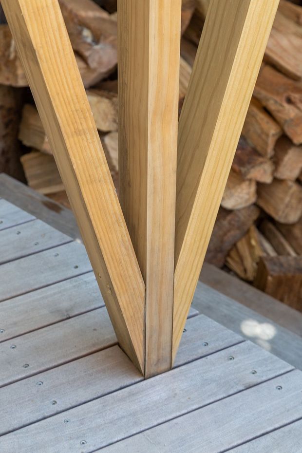 Close-up detail of a timber structural post or angled support set into a weathered grey timber deck. The natural light-wood grain and angled members form a V-shaped junction, with a stacked firewood backdrop visible behind.