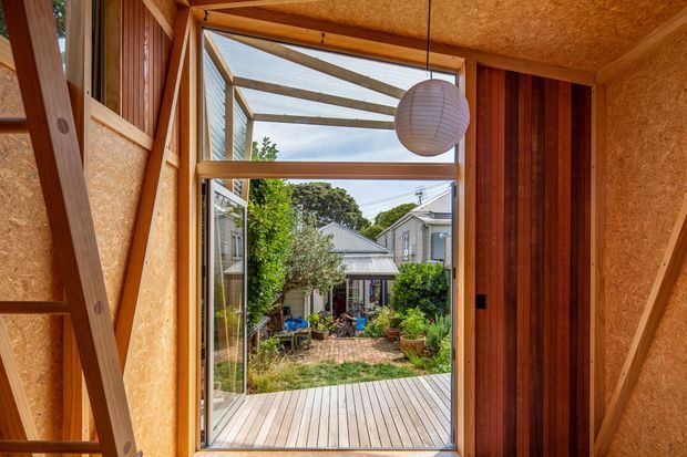 A warm timber-framed interior looking through a large glazed opening onto a small timber deck and lush courtyard garden. The room features exposed structural timber framing, textured wood-fibre or OSB-like wall and ceiling panels, vertical dark timber panelling, and a suspended spherical paper pendant light. A residential courtyard and neighbouring homes are visible beyond.