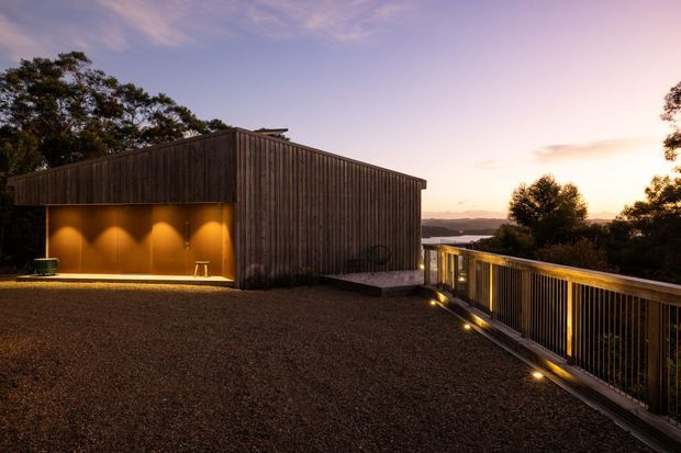 A contemporary timber-clad building viewed at dusk from a broad gravel terrace or rooftop deck. The structure has a simple sloping roof, vertical dark timber cladding, and a warmly illuminated recessed outdoor area. A metal-and-timber balustrade runs along the edge, overlooking a wooded, hilly landscape and distant water.