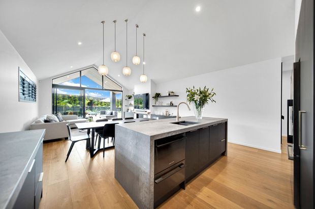 Bright open-plan kitchen and living area with a large dark island, pale stone benchtops, timber flooring, pendant lighting, minimalist white walls, and extensive glazing overlooking greenery. The kitchen opens directly to a dining and lounge space.