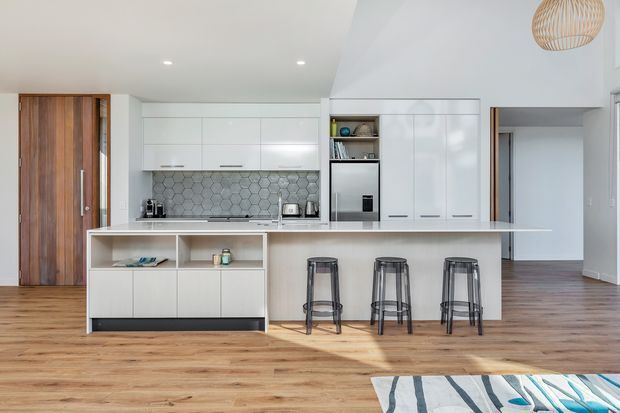 Bright, open-plan contemporary kitchen viewed from the living area, with a large white island and integrated cabinetry, three bar stools, timber flooring, a hexagonal grey tile splashback, and a warm timber entry door. A hallway and adjacent room are visible beyond the kitchen. The space has clean lines, recessed ceiling lights, and a woven pendant light.