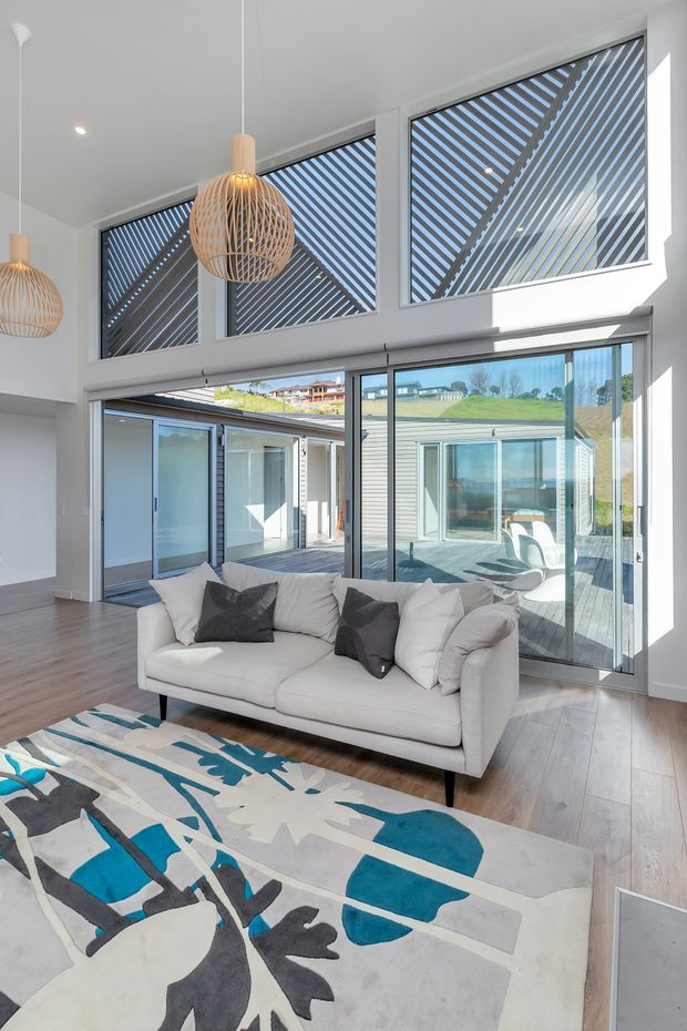 Bright contemporary living room with a white sofa, large abstract area rug, timber flooring, sculptural woven pendant lights, and extensive floor-to-ceiling glazing. High clerestory windows feature diagonal external screening, while sliding glass doors open toward a timber deck and adjoining courtyard-like spaces. The room has an airy, open connection to the outdoors and abundant natural light.