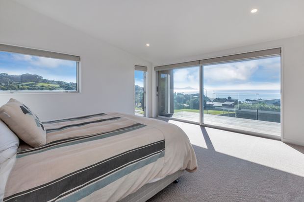 A bright, contemporary bedroom with a low-profile bed dressed in a striped neutral, charcoal, and pale blue duvet. White walls, recessed ceiling lights, and grey carpet create a clean, understated interior. Large corner windows and sliding glass doors provide expansive views toward a green hillside and the ocean, with a balcony or terrace visible outside.
