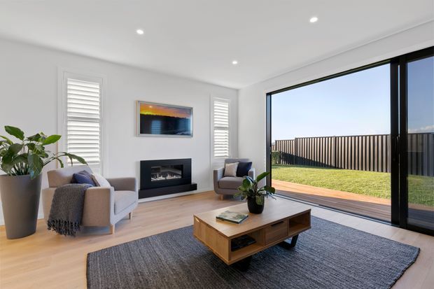 Bright contemporary living room with white walls, light timber flooring, plantation shutters, a black-framed expansive sliding glass door, and a view to a fenced lawn and timber deck. The room includes two upholstered armchairs, a low timber coffee table, a dark textured rug, indoor plants, and a minimalist black fireplace surround beneath framed artwork.