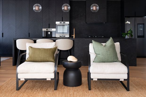 A sophisticated open-plan living room and kitchen with a dark, near-black palette. Two cream upholstered lounge chairs with black timber frames face the camera, separated by a rounded black side table. Behind them is a dark kitchen with a black island, integrated appliances, pendant lights, dark cabinetry and a warm brass tap. A natural woven rug, pale upholstery and muted sage and olive cushions add warmth and contrast.