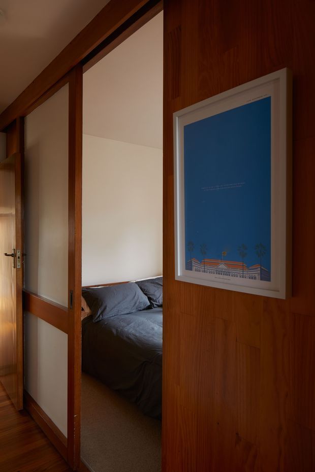A warm, dimly lit interior view through a timber-framed sliding or hinged door into a compact bedroom. The bedroom contains a dark grey bed with rumpled bedding, while the surrounding walls and door are clad in rich vertical timber. A framed blue artwork is mounted on the timber wall in the foreground. The composition emphasises the doorway, timber detailing, and intimate connection between hallway and bedroom.