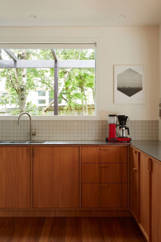 Bright compact kitchen with warm mid-century timber cabinetry, a grey tiled splashback and benchtop, stainless-steel sink and tap, and a large window overlooking leafy greenery. A red coffee machine sits on the benchtop, while framed artwork adds a small decorative element.