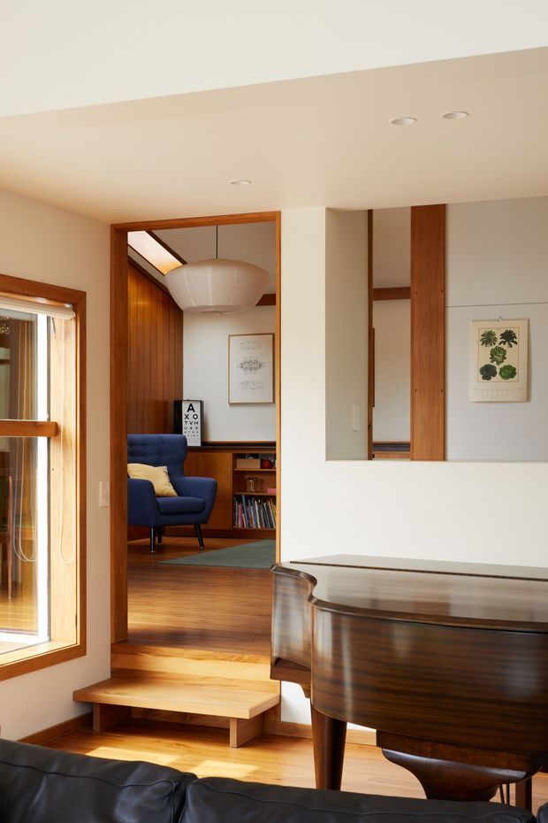 A warm mid-century-inspired interior view from a living area toward a slightly elevated study or sitting room. The foreground contains a dark timber baby grand piano and part of a black leather sofa. A short timber step leads to the adjoining room, which features rich wood wall panelling, built-in shelving, a blue lounge chair, framed artwork, and a large sculptural pendant light. Timber-framed glazing and recessed ceiling lights complete the space.