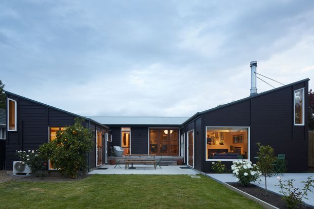 Single-storey U-shaped contemporary house viewed from the landscaped front yard. The dark horizontal-clad wings frame a central recessed entry courtyard with timber-framed glazing and warm interior lighting. Large windows reveal a kitchen or living area, while a covered or flat-roofed connecting section links the wings. The scene includes lawn, paving, garden planting, and a visible metal chimney.