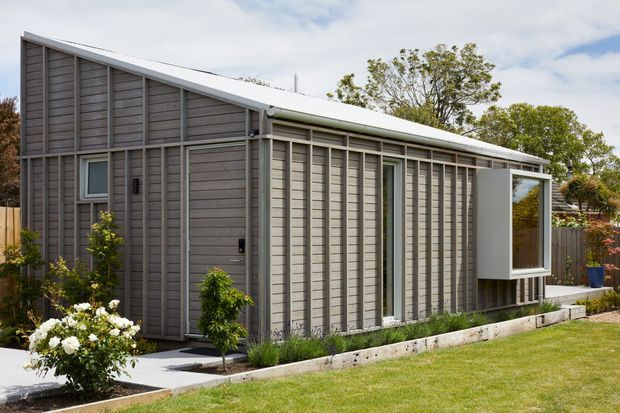 A compact single-storey contemporary dwelling with a steep asymmetrical pitched roof and extensive grey horizontal weatherboard cladding. The facade features tall narrow windows, a projecting glazed box window, white trim, a simple concrete-edged garden bed, and a neat lawn with established planting.