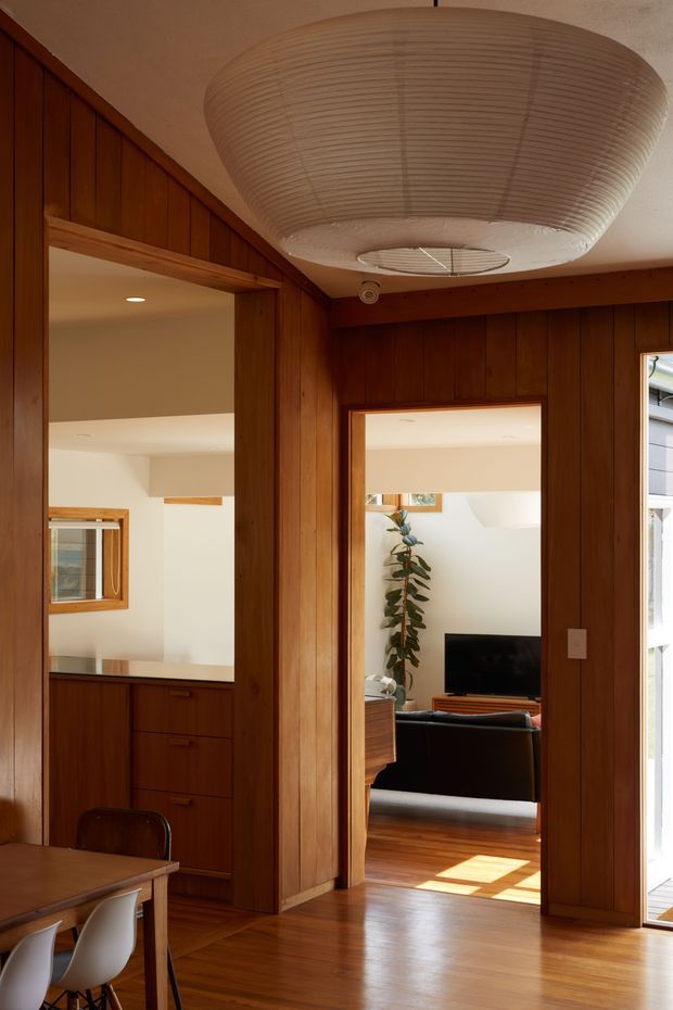 Warm interior view of a mid-century-inspired timber-lined circulation and dining area. Dark natural wood vertical wall panelling and matching timber floors frame openings into a compact kitchen and living room. A large sculptural ribbed ceiling light dominates the space, while simple dining furniture, built-in cabinetry, and a television seating area are visible beyond.