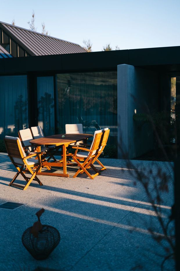 A contemporary outdoor patio or terrace beside a dark, low-profile building. The space features a timber dining table with folding wooden chairs, large floor-to-ceiling glazed panels, a smooth concrete column, dark cladding, and a pitched metal roof visible behind the facade. Strong low-angle sunlight creates long shadows across the paved terrace.