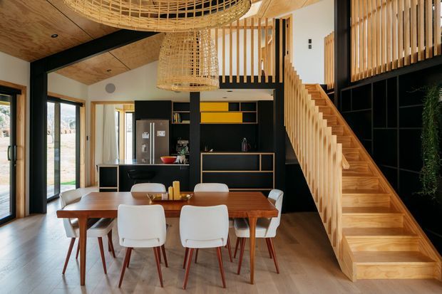 Warm contemporary open-plan dining area with a timber dining table and white upholstered chairs, adjoining a black built-in kitchen and storage wall. A timber staircase with vertical slatted balustrades rises to a mezzanine, while large glazed doors bring in natural light. The room features timber flooring and ceiling panels, black joinery, yellow accent shelving, and woven pendant lights.