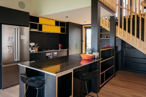 A contemporary open-plan kitchen with a large dark island bench, black built-in cabinetry, stainless-steel appliances, and striking mustard-yellow accent cupboards. Black wire bar stools sit at the island, while a timber staircase with vertical balustrades rises beside dark built-in shelving. Warm timber flooring and daylight create a refined, high-contrast interior.