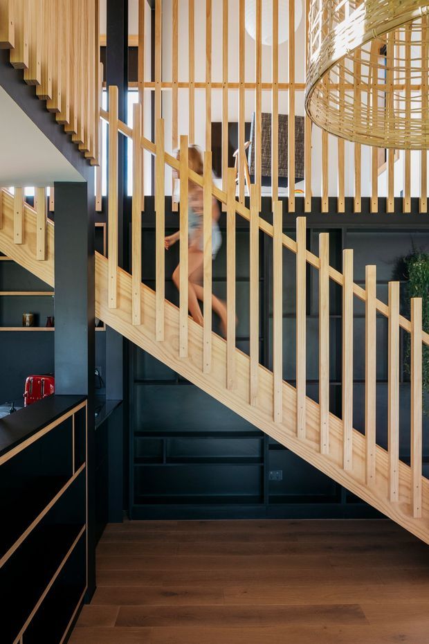 A contemporary interior stairway with a sculptural light timber staircase and vertical timber balustrade slats. The stairs pass through a dark charcoal built-in joinery and wall backdrop, with warm dark timber flooring, tall glazing, and a large woven pendant light creating a strong contrast between natural wood and deep charcoal surfaces.
