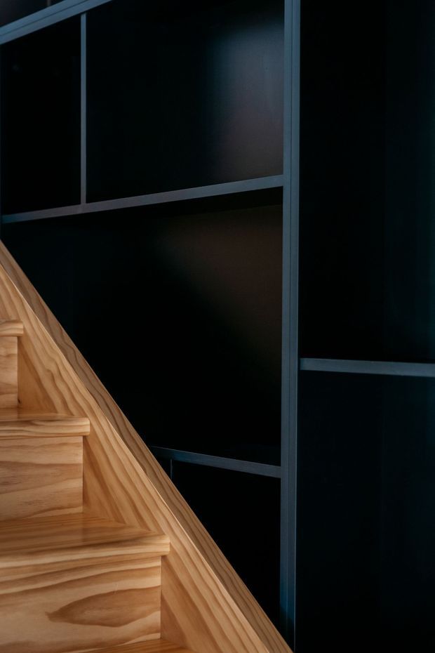 Close-up architectural detail of a timber staircase with prominent natural grain, set against a dark black wall or panelled backdrop with slim dark framing. The image emphasizes the contrast between warm wood and the moody dark surround.