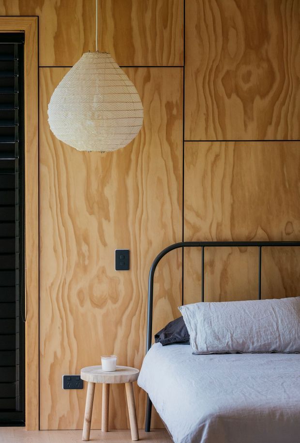A warm, tactile bedroom interior featuring full-height natural plywood wall panelling with visible grain and dark joint lines. A black metal-framed bed with pale linen bedding sits beside a small timber stool table, while a woven pendant light hangs against the timber wall. A dark louvred window or door is visible at left.