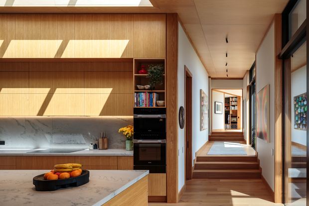 A warm, light-filled contemporary kitchen opening onto a long residential hallway. The kitchen features vertically grained timber cabinetry, a pale veined stone benchtop and backsplash, integrated dark appliances, open shelving, and a central island. The hallway has timber flooring, timber-lined ceilings, framed artwork, glazed openings, and a short set of steps leading toward a built-in bookcase and another room. Strong sunlight enters through skylights or clerestory glazing, creating geometric patches of light.