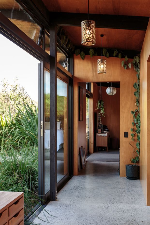 A warm, timber-lined interior passage or entry corridor with full-height dark-framed glazing on one side, opening onto dense planting. The space features a polished concrete floor, rich wood wall and ceiling panels, dark structural beams and trims, hanging decorative pendant lights, and trailing indoor plants. A bedroom is partially visible through the glazing, with additional rooms extending down the corridor.