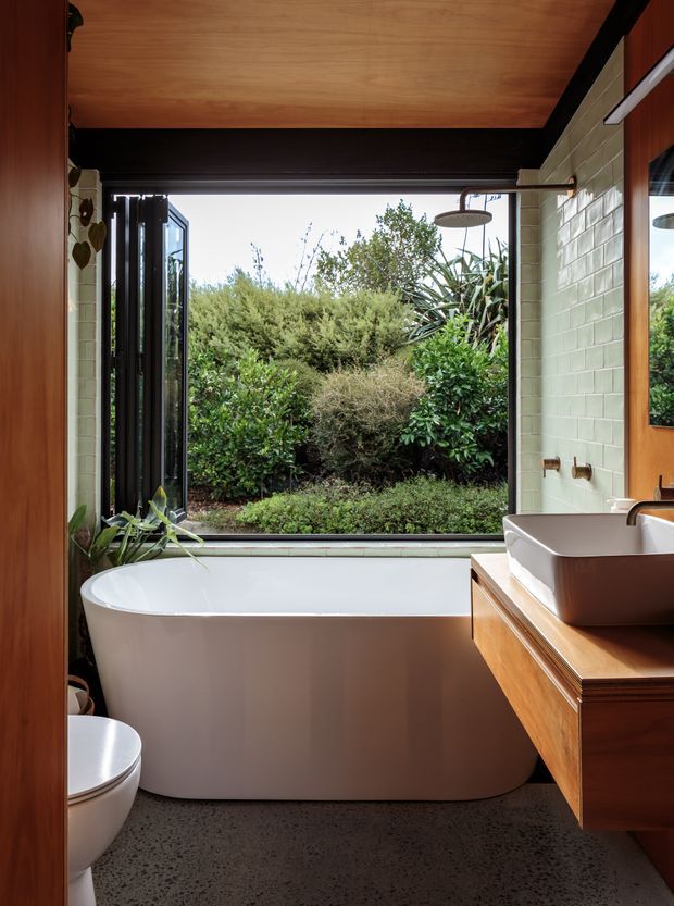 A compact, contemporary bathroom with a freestanding white bathtub positioned beneath a large opening to a lush garden. The room features pale green glazed wall tiles, a dark structural beam, warm timber ceiling and cabinetry, a white vessel basin, wall-mounted taps, and a terrazzo-like grey floor. A toilet is partially visible at the left edge, while a black-framed window or door opens toward the planted exterior.