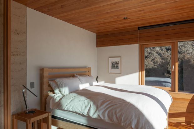 A warm, compact bedroom with a timber platform bed, white bedding, integrated bedside lighting, and a full-width timber-framed window overlooking trees. The room features a rich timber slatted ceiling, pale plaster walls, natural stone or concrete wall detailing, and built-in timber window seating. Strong afternoon sunlight creates a calm, natural atmosphere.