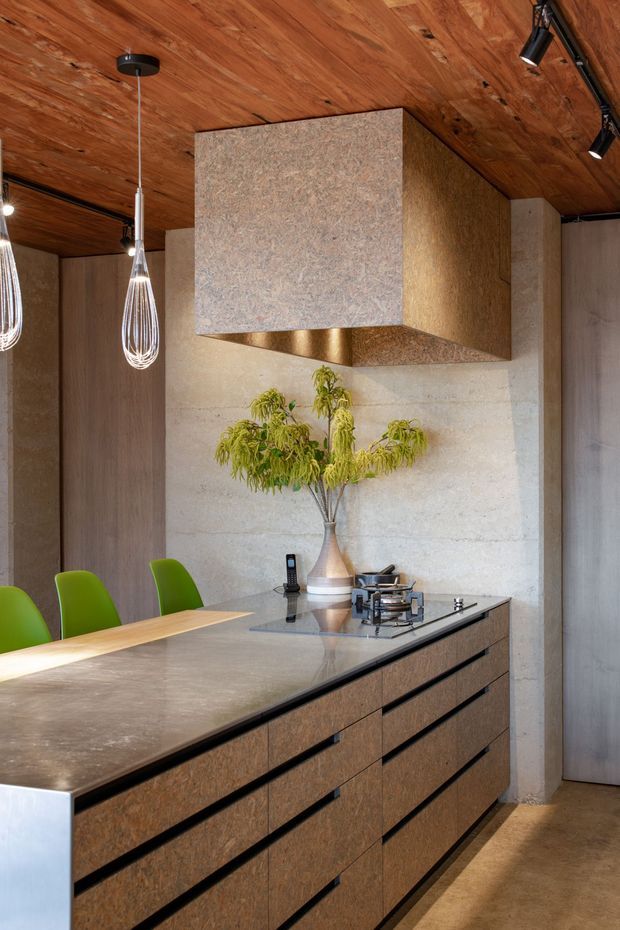 A sophisticated contemporary kitchen centered on a large island with a grey stone benchtop, dark wood-toned cabinetry, integrated gas cooktop, and black edge detailing. A textured stone or concrete feature wall supports a large sculptural rangehood clad in a warm metallic finish. The ceiling is lined with warm timber boards, while green dining chairs, pendant lights, and a natural branch arrangement add contrast and character.