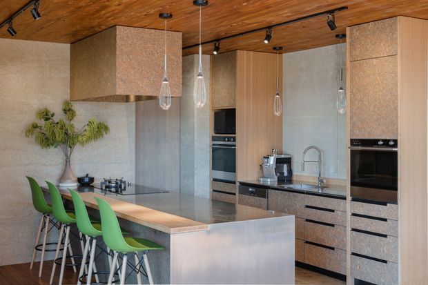 Contemporary kitchen with a large island and integrated green bar stools. The space features warm timber ceiling lining, concrete-look wall surfaces, extensive built-in cabinetry, stainless steel appliances, a stone or engineered-stone island benchtop, and sculptural pendant lights.