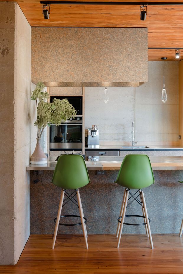 A compact contemporary kitchen centred on a peninsula breakfast bar with two green bar stools. The space features warm timber flooring and a timber-lined ceiling, textured stone-like cabinetry and rangehood surround, pale concrete-look walls, stainless-steel appliances, and suspended pendant lights. A large vase with foliage adds a natural accent.