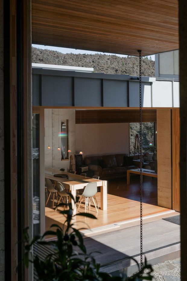 View through a large open glazed opening into a warm contemporary open-plan living and dining area. The space features extensive timber lining on the ceiling and floor, exposed concrete or stone-toned walls, a dark leather sofa, timber dining table with light chairs, and strong indoor-outdoor connection. A dark metal overhead structure and rain chain are visible, with a wooded landscape beyond.
