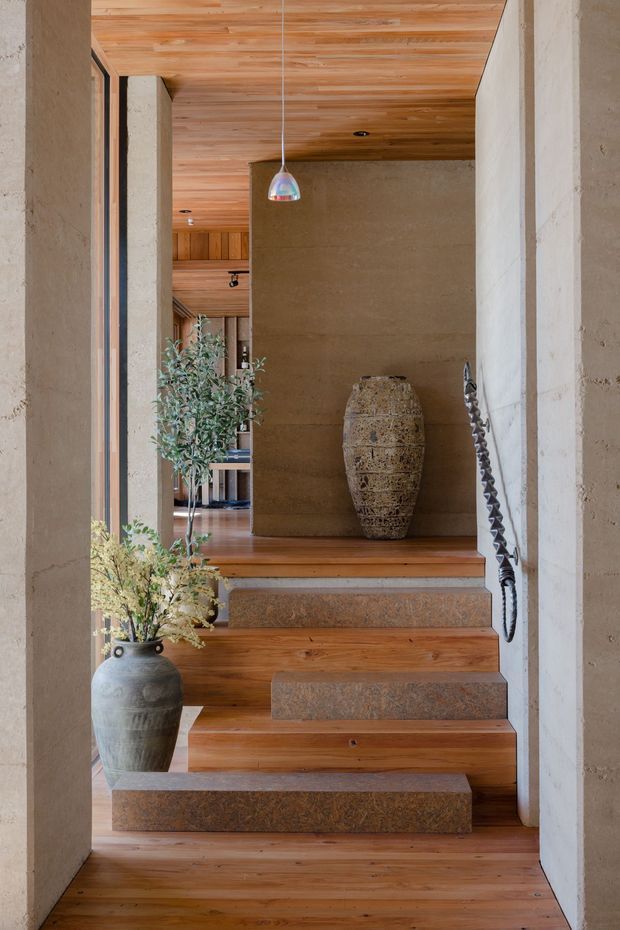 A warm, sculptural interior entry sequence with floating timber stair treads and thick stone or concrete risers leading to a raised landing. The space features textured earthen-toned walls and substantial columns, a timber-lined ceiling and floor, narrow dark-framed glazing, a suspended pendant light, a large decorative vessel, and indoor plants. A dark sculptural handrail follows the stair on the right.