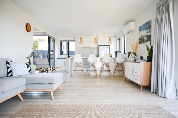 Bright open-plan coastal-style living, dining, and kitchen interior with pale timber-look flooring, white walls and cabinetry, a light grey sectional sofa, timber-legged dining chairs, pendant lights, indoor plants, and full-height curtains. Large windows provide abundant natural light, and a low timber sideboard sits along the right wall.