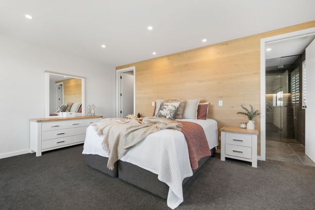 Bright contemporary bedroom with a large bed centered against a full-height warm timber feature wall. The room has dark grey carpet, white walls and ceiling with recessed downlights, white bedside drawers, and a matching white dresser with mirror. A glazed or open doorway at right reveals an adjoining tiled bathroom with dark-framed glazing and a shower.