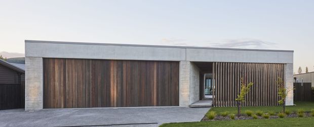 Front exterior view of a low, single-storey contemporary residence with a flat roof, broad vertical dark timber garage doors, a matching timber privacy screen, pale concrete or rendered masonry walls, and a recessed glazed entry. The house sits behind a simple paved driveway and narrow landscaped garden.