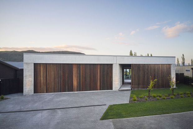 A low, contemporary residential frontage with a broad flat-roofed concrete garage volume, warm vertical timber garage doors and a matching timber privacy screen. A recessed pedestrian entry divides the garage and screened area, opening toward a small landscaped lawn and concrete driveway. The scene is photographed from outside in soft evening light, with a suburban landscape in the background.