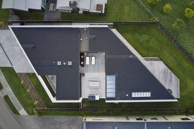 Aerial top-down view of a contemporary residential building, dominated by dark metal pitched roof planes arranged around a central light-coloured roof terrace or courtyard zone. Several skylights and roof penetrations are visible, along with a long linear rooflight. The property sits within a landscaped suburban setting with lawns, planted edges, paths, fencing, and neighbouring buildings.