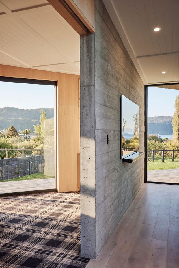 Interior architectural view of a contemporary open-plan living area divided by a substantial exposed concrete feature wall with a wall-mounted television. The space has full-height black-framed glazing opening to a deck and landscaped yard with mountain and lake views. One side features warm timber wall panelling and patterned carpet, while the adjoining area has pale timber flooring. A white paneled ceiling with recessed downlights and exposed timber beams completes the design.