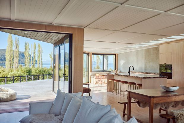 Bright open-plan contemporary living, dining and kitchen interior with extensive glazing opening onto a covered terrace and views of a lake and surrounding landscape. The room features pale timber wall and ceiling finishes, a concrete-look kitchen wall, timber cabinetry, a stone island benchtop with bar seating, and soft neutral furnishings.