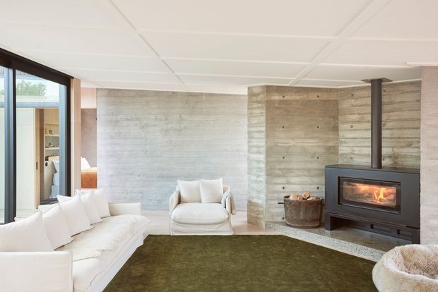 Bright contemporary living room with a white sectional sofa and armchair arranged around a dark green rug, a freestanding wood-burning fireplace, and textured raw concrete walls. Floor-to-ceiling glazing and timber-framed doors bring in natural light, while a white panelled ceiling and concrete structural column reinforce the minimalist architectural character.