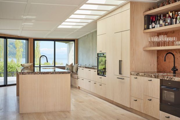 A bright contemporary kitchen with a large light-wood island, extensive floor-to-ceiling cabinetry, integrated appliances, and dark stone benchtops. A built-in open bar and glassware shelving occupy the right side, while large glazed doors and windows connect the kitchen to a leafy outdoor setting. A skylit or illuminated panel ceiling and pale timber flooring add to the airy feel.